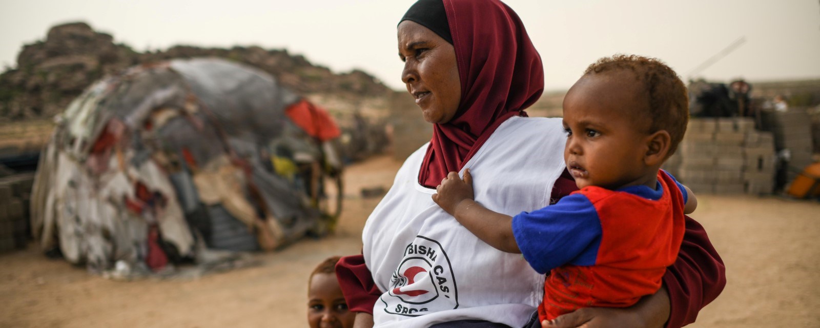 En kvinne i Somalia med Røde Kors-vest bærer et lite barn samtidig som hun holder et annet barn i hånden