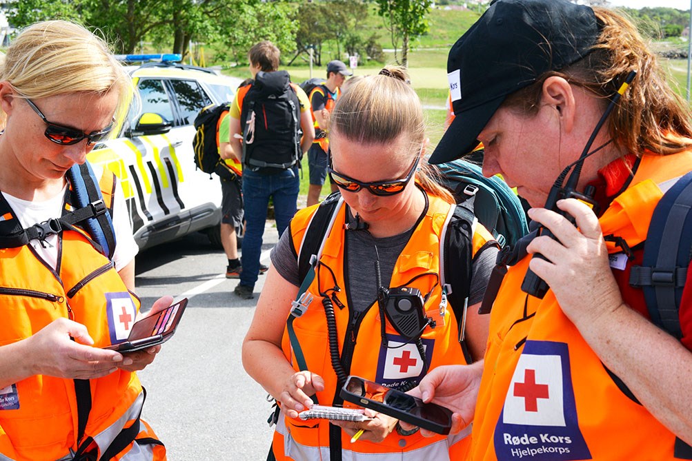 Tre kvinner i Røde Kors-vester ser på mobiltelefon.