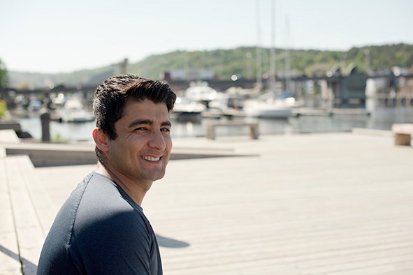 En ung mann sitter i sola ved en båthavn og snur seg mot kamera og smiler.