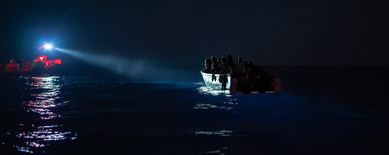 En redningsbåt lyser opp en båt full av mennesker i mørket i Middelhavet.