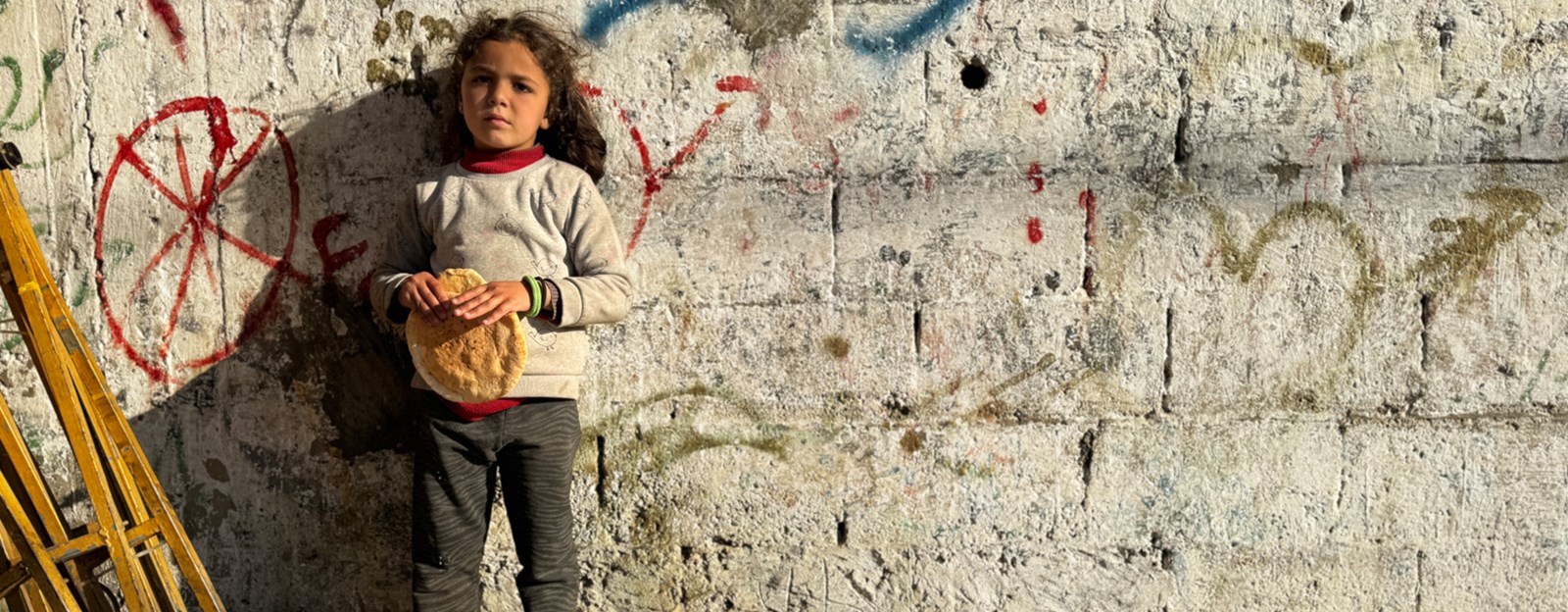 Et barn står inntil en murvegg, ser i kamera og holder brød i hendene